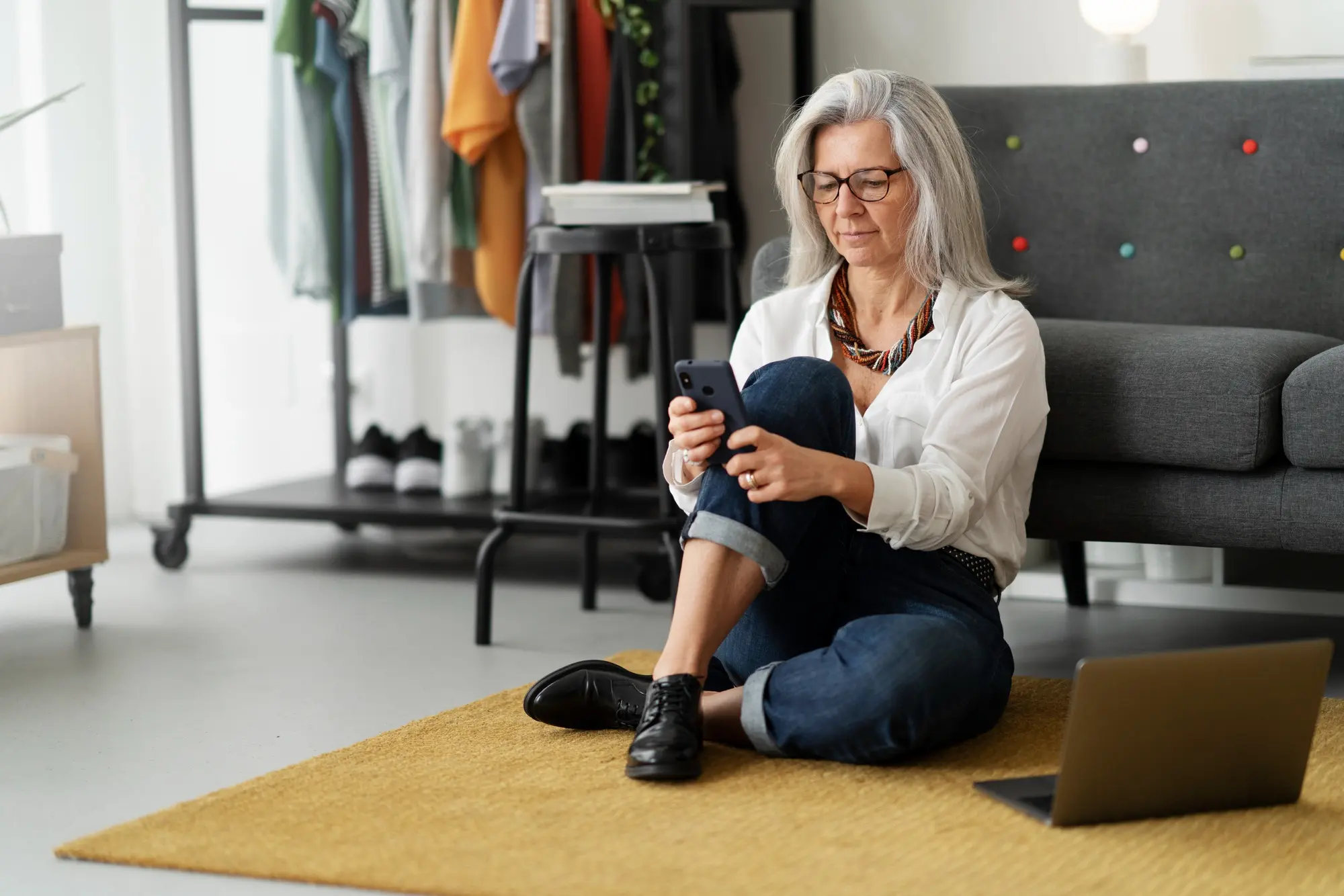 Woman in her early 60s with long grey hair and glasses sitting relaxed on a mustard-yellow rug on the floor, leaning against a grey sofa and looking at her smartphone. White blouse, dark jeans, a laptop beside her. Modern, bright living room with a clothing rack in the background.