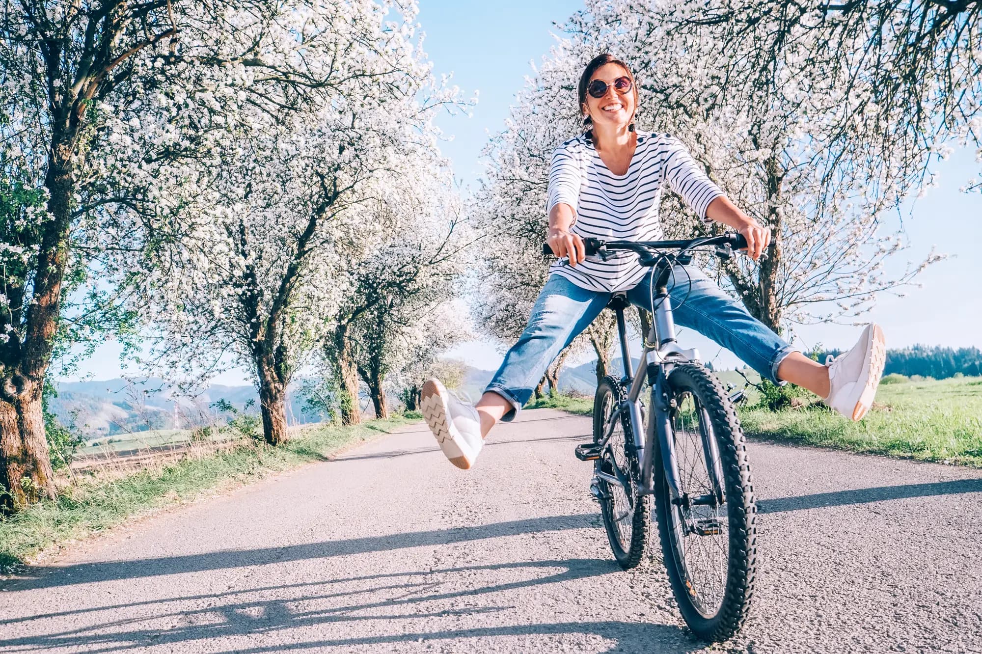 Eine lachende Frau mittleren Alters fährt mit dem Fahrrad eine sonnige Landstraße entlang, gesäumt von weiß blühenden Bäumen. Sie trägt ein Streifenshirt, eine Jeans und eine Sonnenbrille und streckt vor Lebensfreude beide Beine zur Seite aus. Helles Frühlingslicht, unbeschwerte Stimmung.