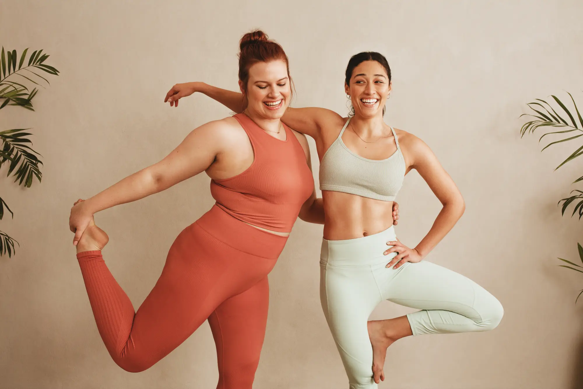 Two laughing women in activewear doing yoga together in front of a warm beige wall. One holds a tree pose, the other balances on one leg. Green houseplants to the sides, a relaxed and empowering atmosphere.