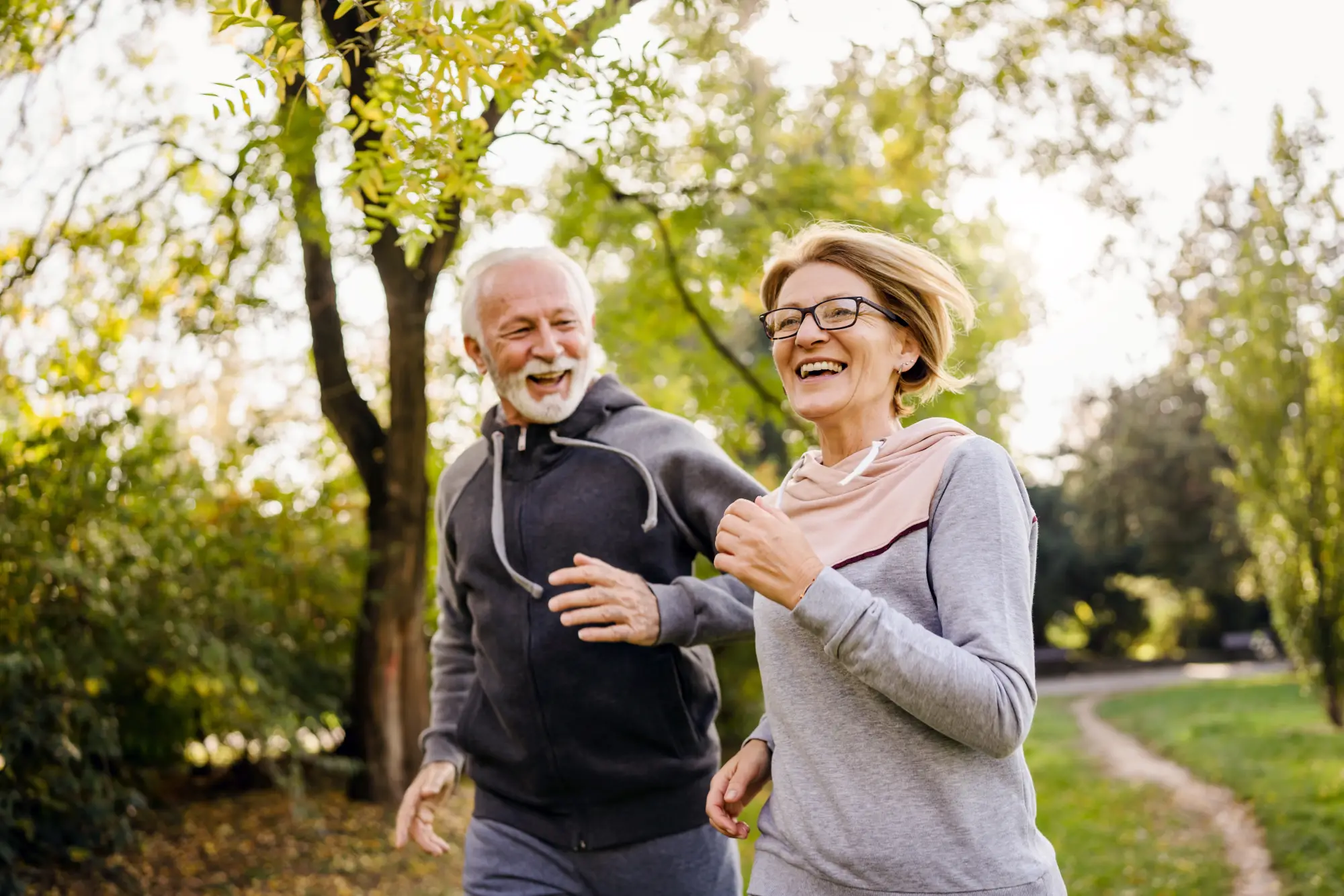 A laughing couple in their early 60s jogging together through a park on a sunny autumn day. Both wear sporty hoodies, with green trees and a winding path in the background. Lively, active mood.