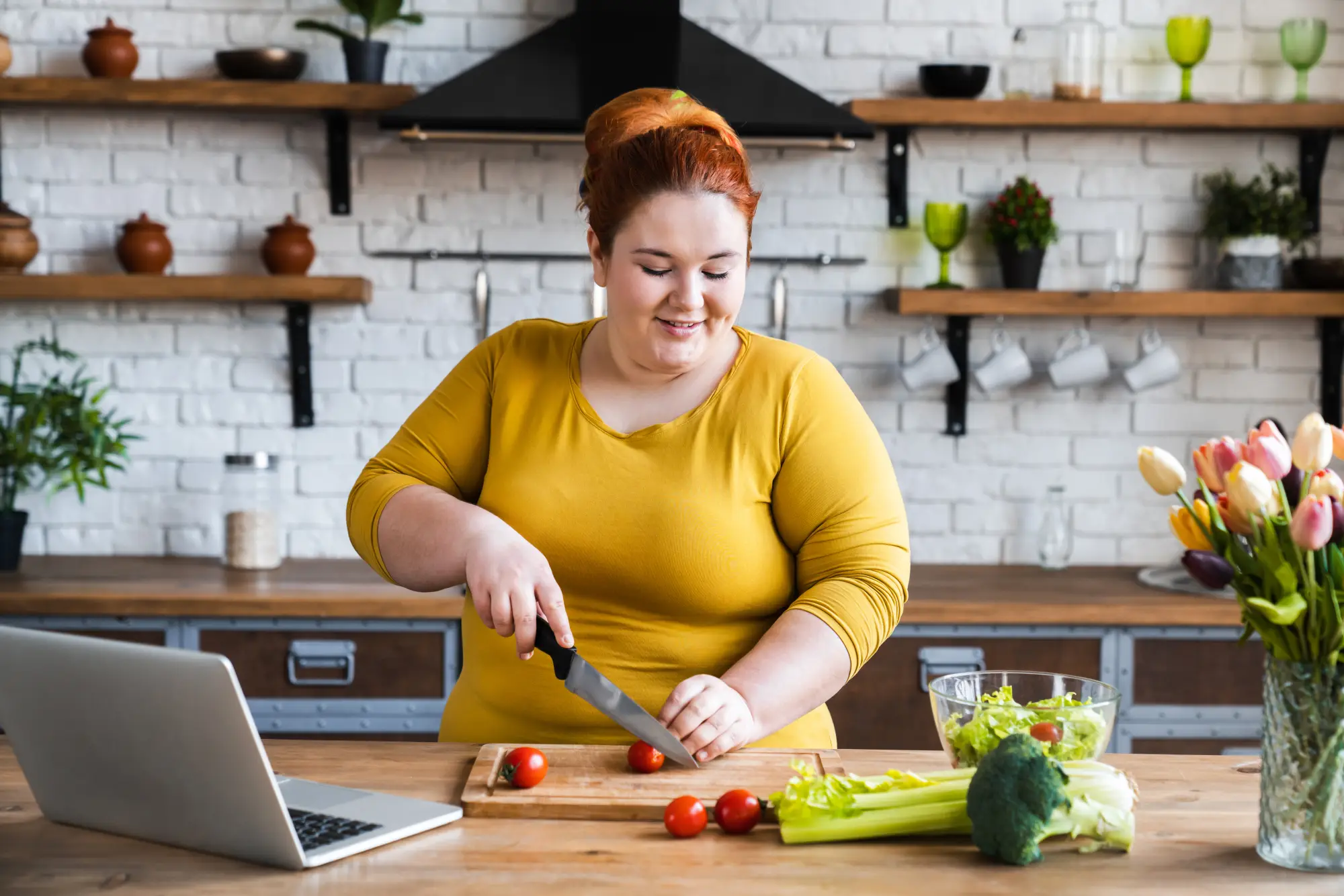 A smiling woman with red hair slicing fresh tomatoes on a wooden board in a bright kitchen. An open laptop sits in front of her, alongside lettuce, celery and broccoli and a bouquet of tulips. Relaxed, healthy everyday mood.