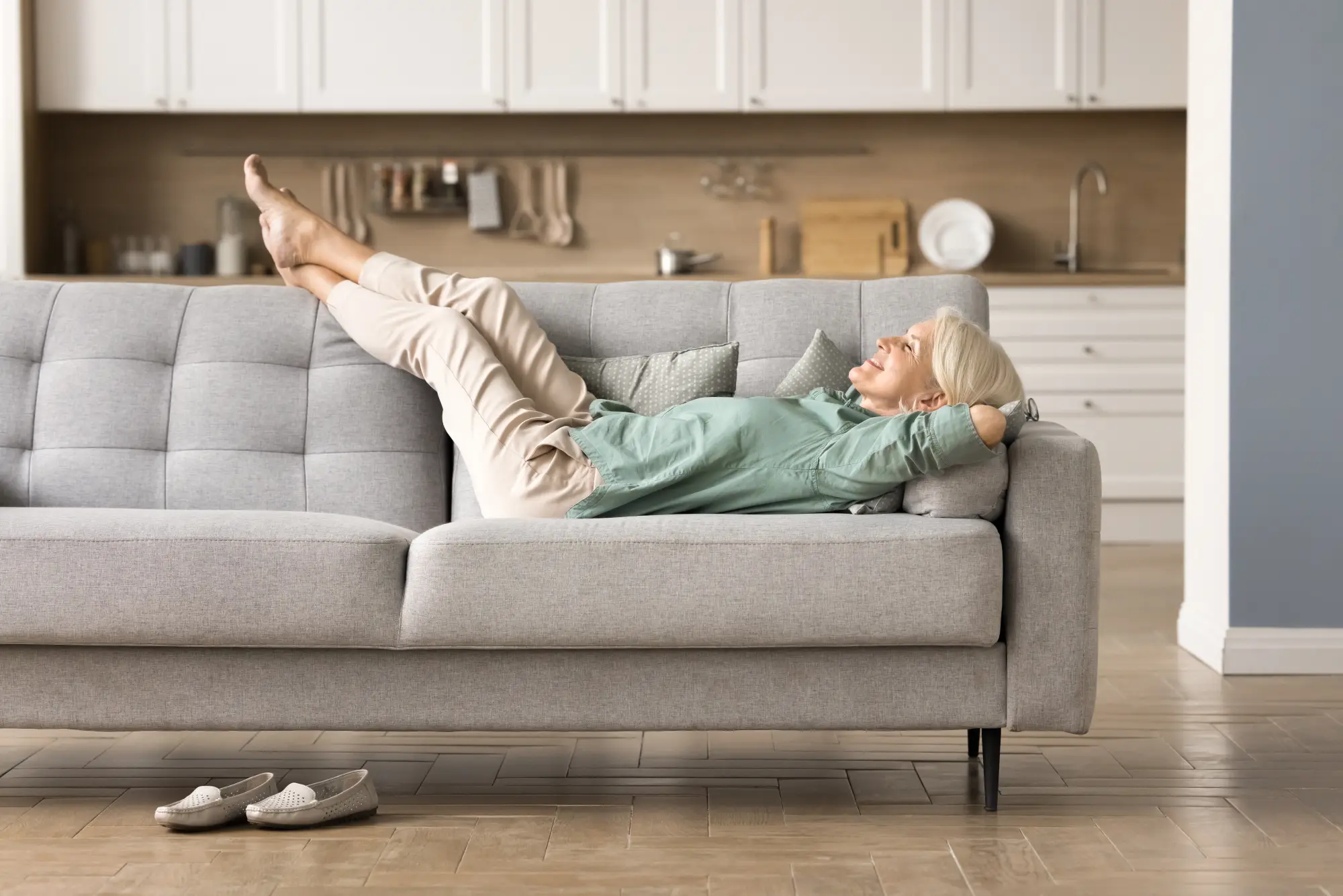 Woman in her early 60s with silver-grey hair relaxing on a grey sofa, legs propped up and hands clasped behind her head. Green blouse, cream trousers, barefoot, with loafers on the floor by the sofa. A bright open kitchen in the background, calm afternoon mood.